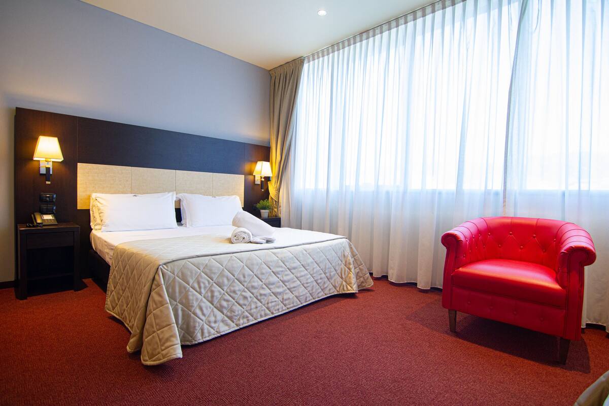 Quarto de hotel com uma cama grande, duas luminárias de cabeceira, uma poltrona vermelha e uma janela com cortinas leves.