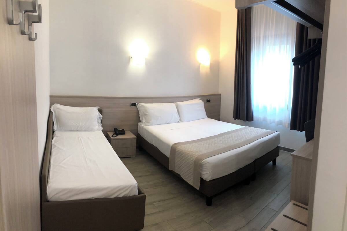 A hotel room with two single beds, a nightstand with a phone, a headboard, and a window with curtains.