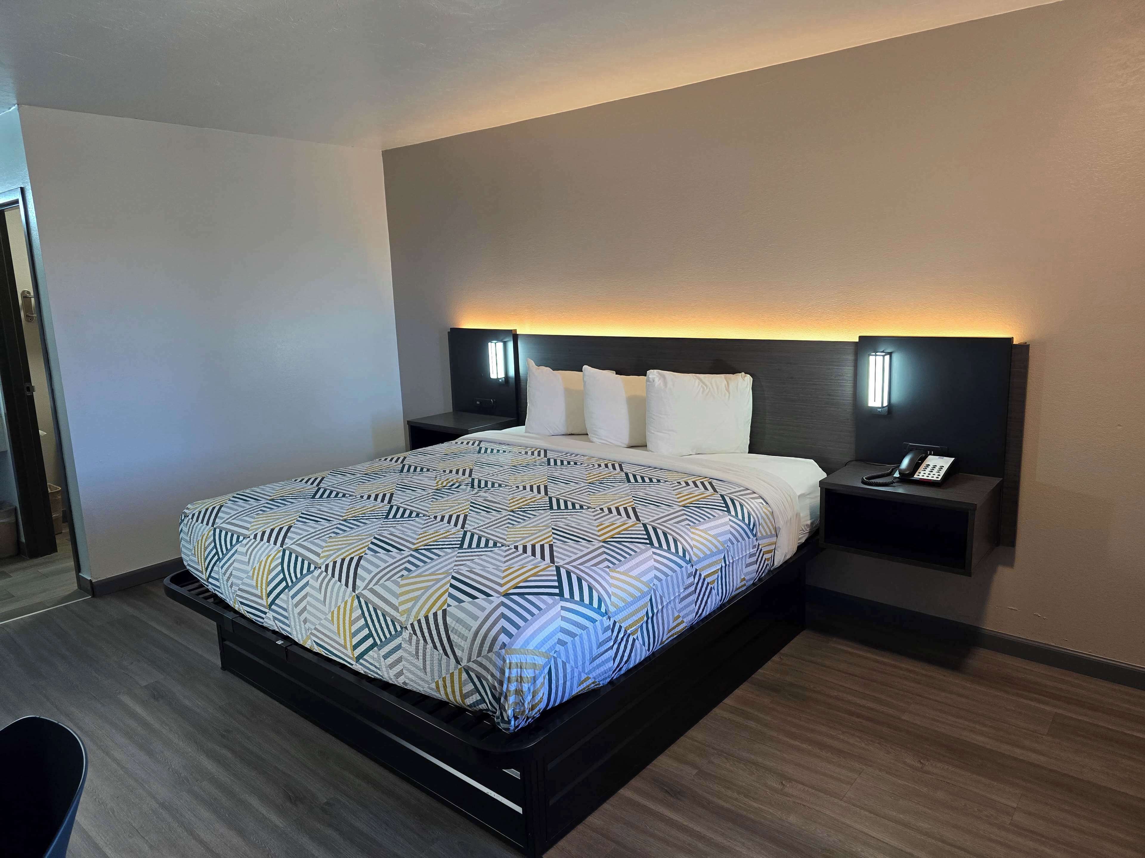A modern hotel room with a large bed, a bedside table with a phone, and a wall-mounted light.