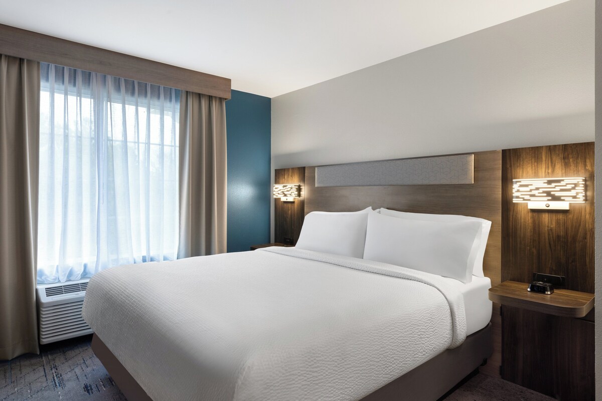 A hotel room with a large bed, a nightstand, a wall-mounted lamp, and a window with curtains.