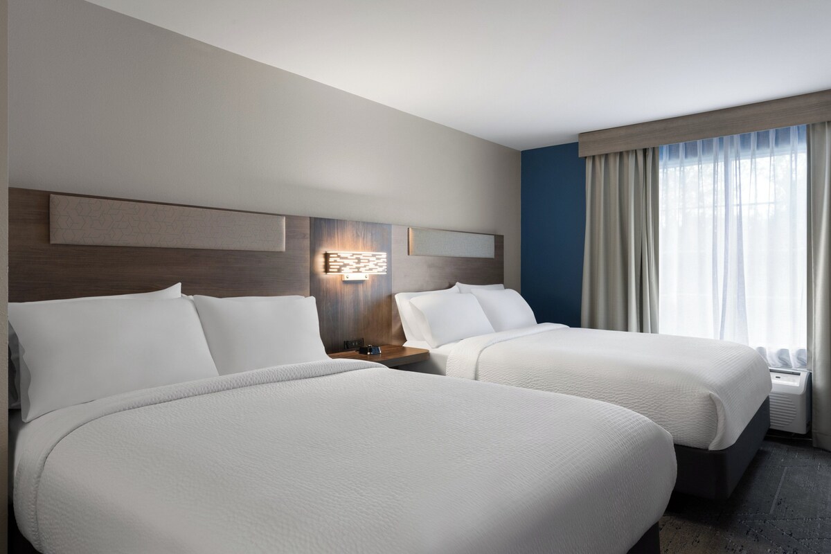 A hotel room with two beds, a nightstand, a wall-mounted light, and a window with curtains.