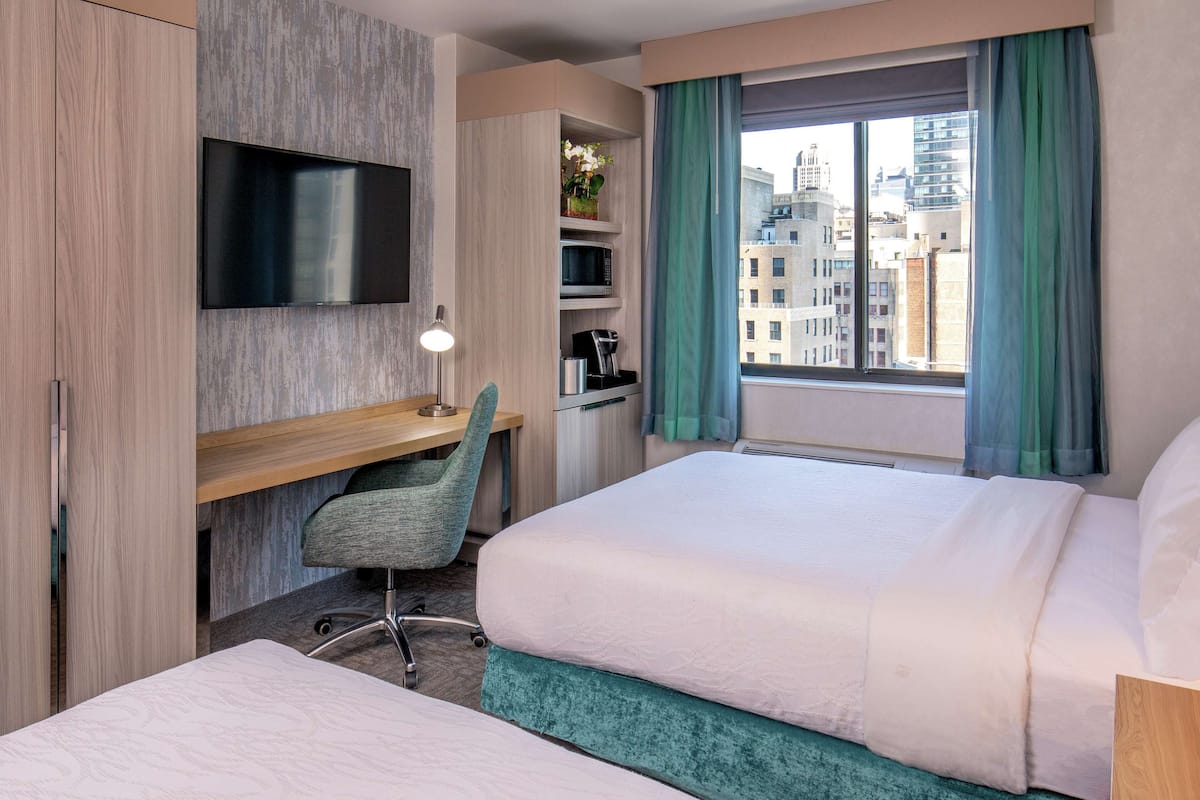 Une chambre d’hôtel comprenant un lit, un bureau avec une chaise, une télévision, une machine à café et une fenêtre avec des rideaux.