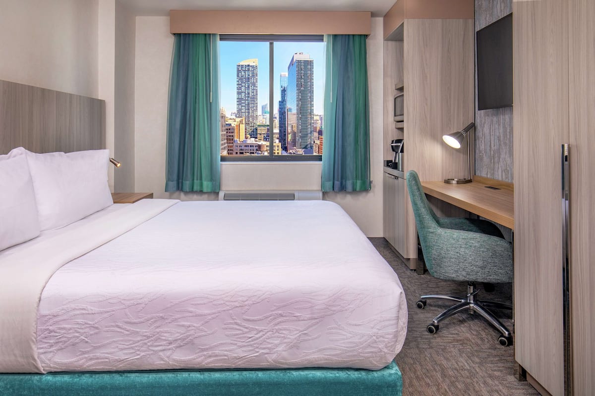 Une chambre d’hôtel avec un grand lit, un bureau avec une chaise, une vue sur la ville et un téléviseur fixé au mur.