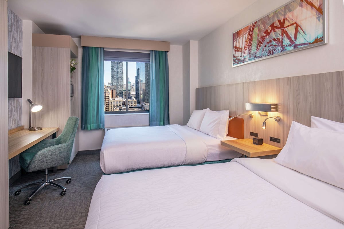 Hotelkamer met twee bedden, een bureau, een stoel, bureaulamp, nachtkastje met lamp, raam met gordijnen en uitzicht op de stad.