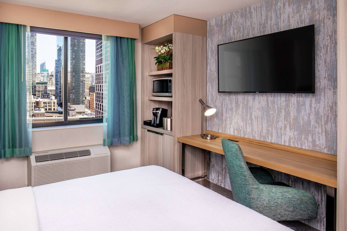 Une chambre d’hôtel comprenant un lit, un bureau avec une chaise, un téléviseur à écran plat, une fenêtre avec des rideaux, une étagère avec un micro-ondes et une plante, et une vue sur la ville.