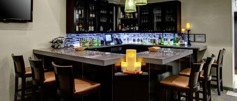 Zona de bar con una isla central, taburetes de bar, televisor de pantalla plana y una variedad de botellas y vasos.