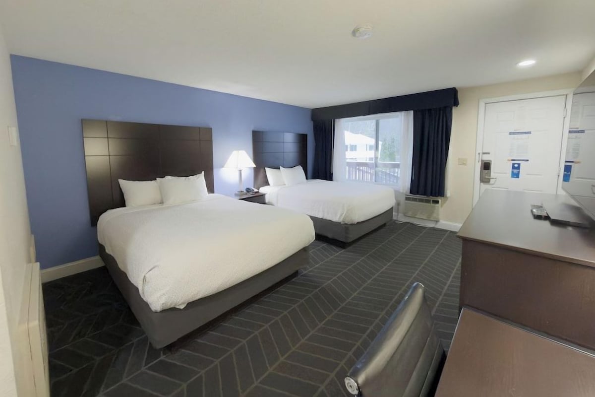 A hotel room with two beds, a desk, and a view of the outside.