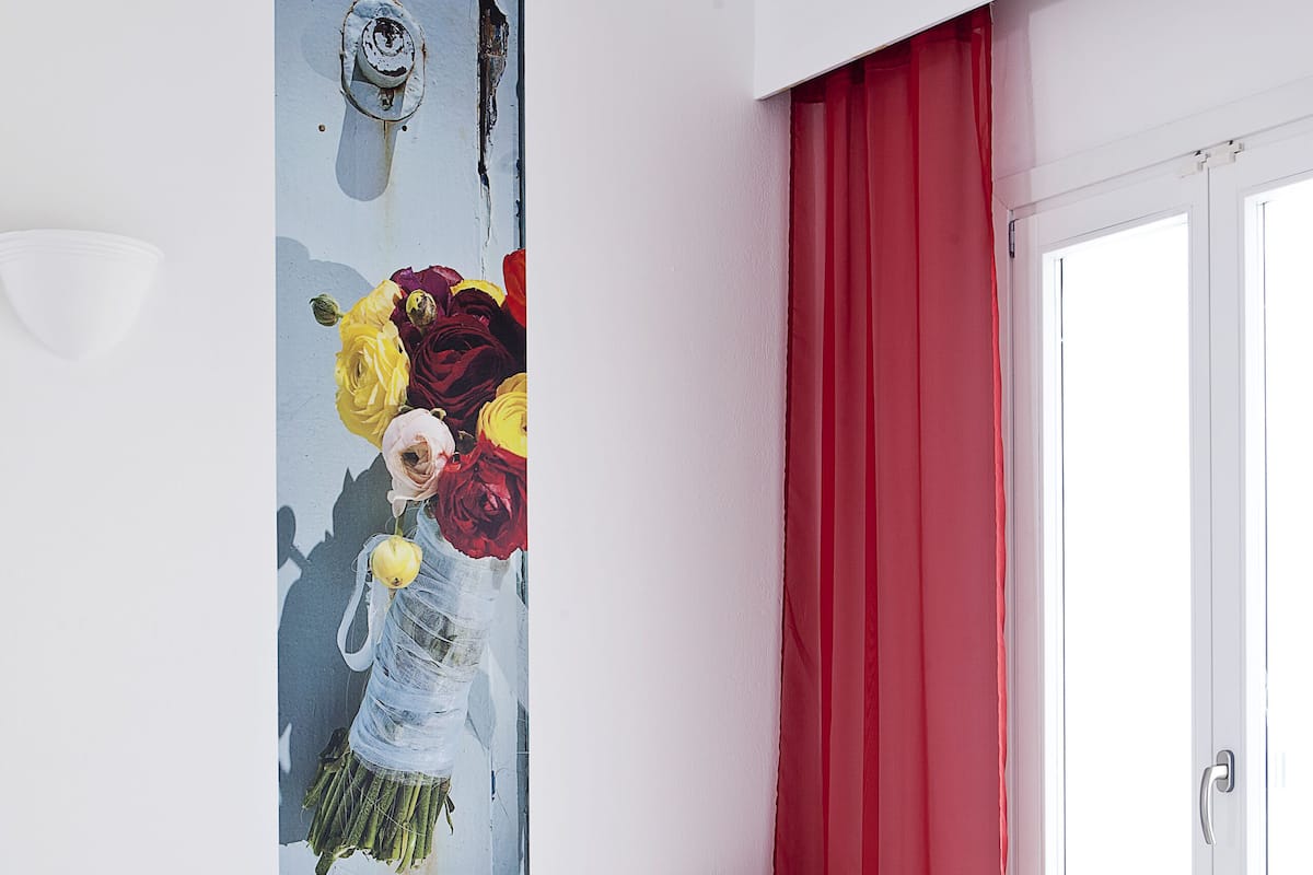 Une chambre d’hôtel avec un lit, deux chaises blanches, une petite table et un tableau représentant des fleurs accroché au mur.