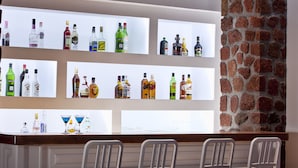 Un espace bar avec plusieurs étagères remplies de bouteilles d’alcool, un mur en briques et quatre chaises blanches.