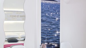 Une chambre à coucher avec un lit, deux chaises blanches, une petite table ronde sur laquelle se trouve un livre, et un miroir mural qui reflète un paysage pittoresque sur l’eau.