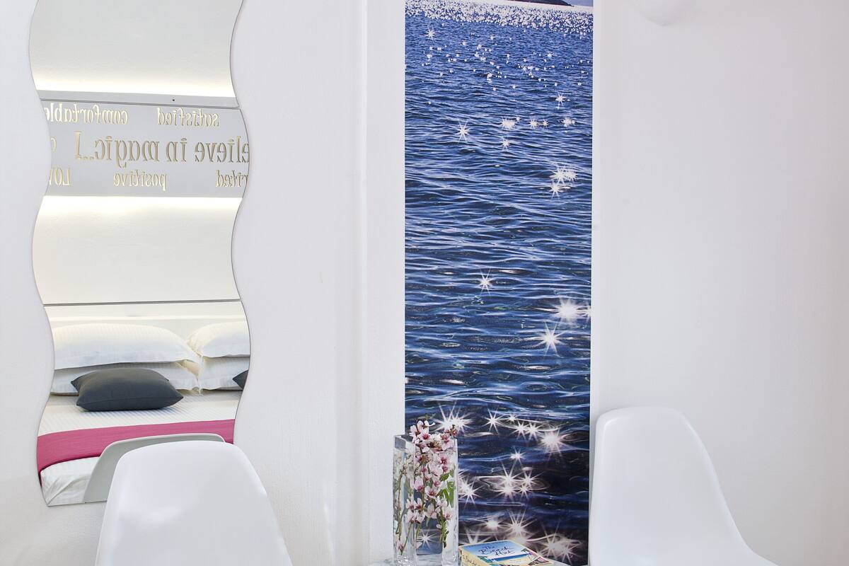 Une chambre à coucher avec un lit, deux chaises blanches, une petite table ronde sur laquelle se trouve un livre, et un miroir mural qui reflète un paysage pittoresque sur l’eau.