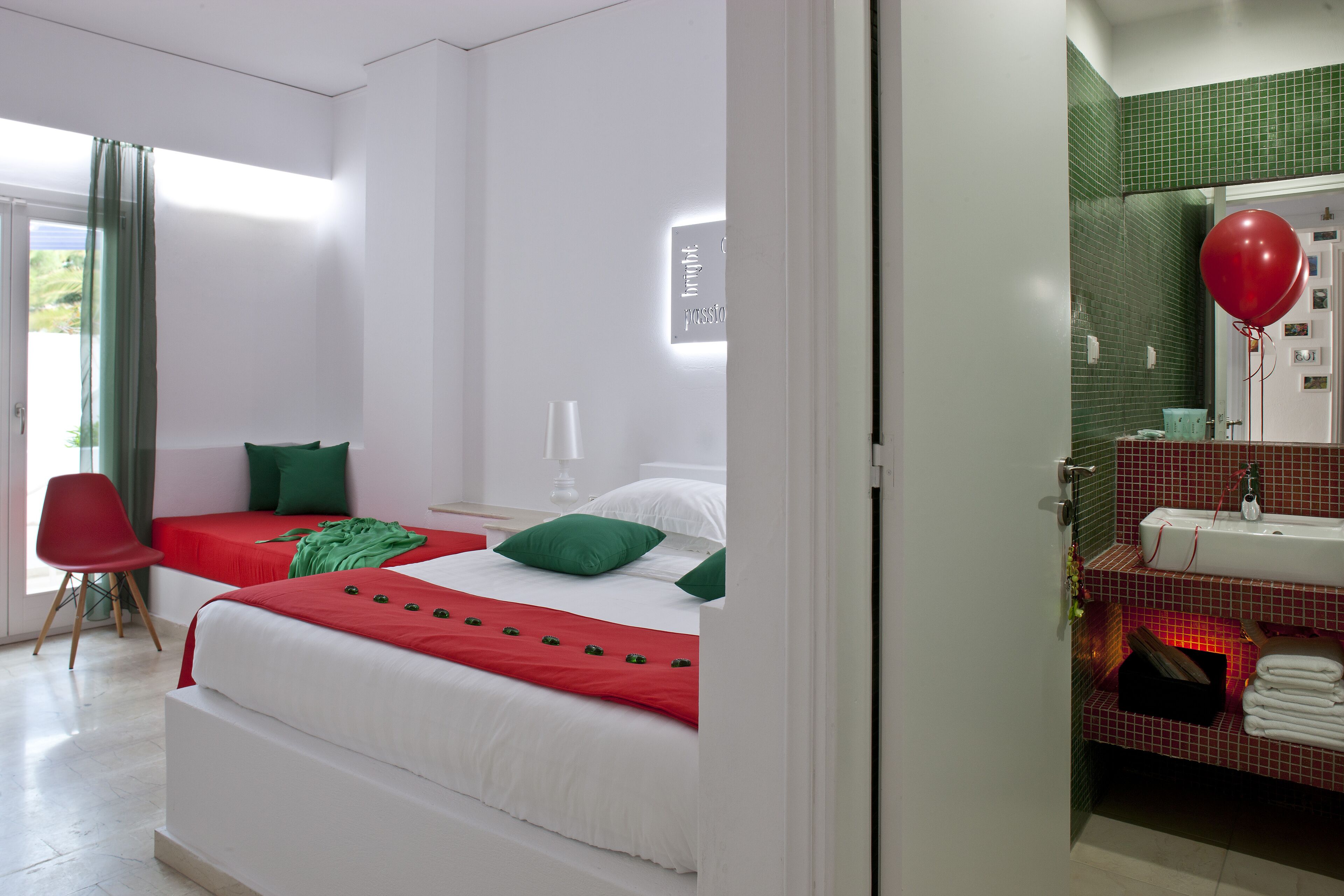 A hotel room with two beds, a red chair, a bathroom with a sink and mirror, and a green balloon.