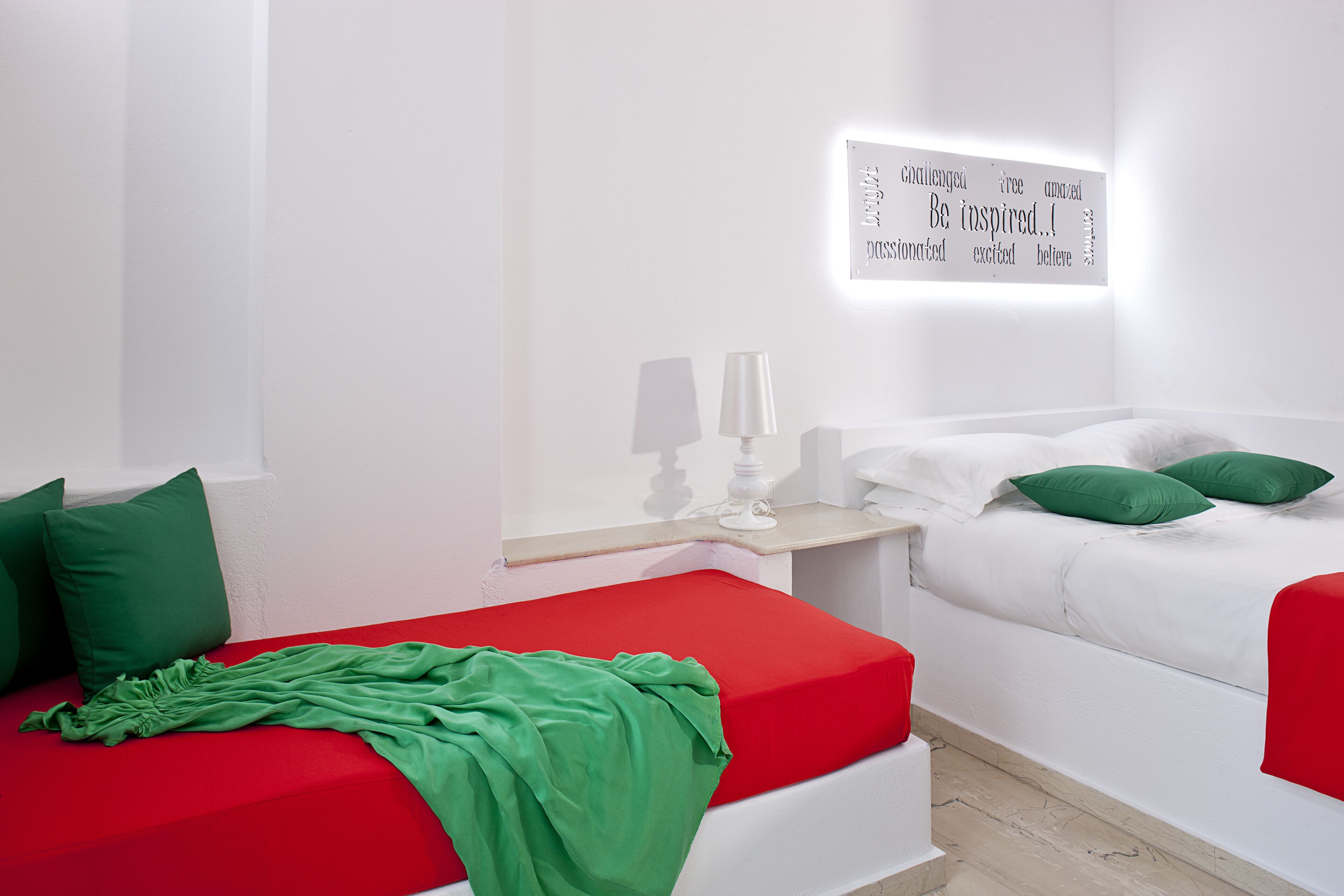 A modern bedroom with two beds, one with a red bedspread and the other with a white bedspread. There is a wall-mounted light fixture with inspirational words.