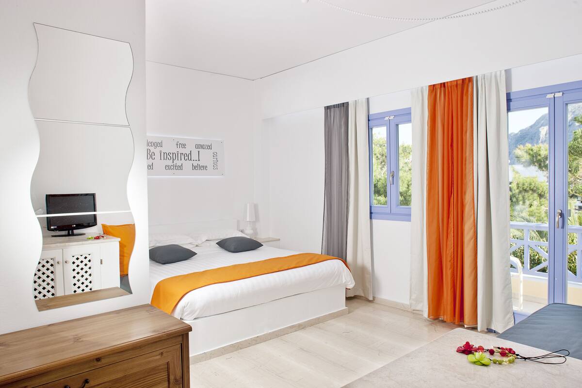 Une chambre à coucher avec un lit, un téléviseur posé sur un meuble, une commode en bois et une vue sur l’extérieur.