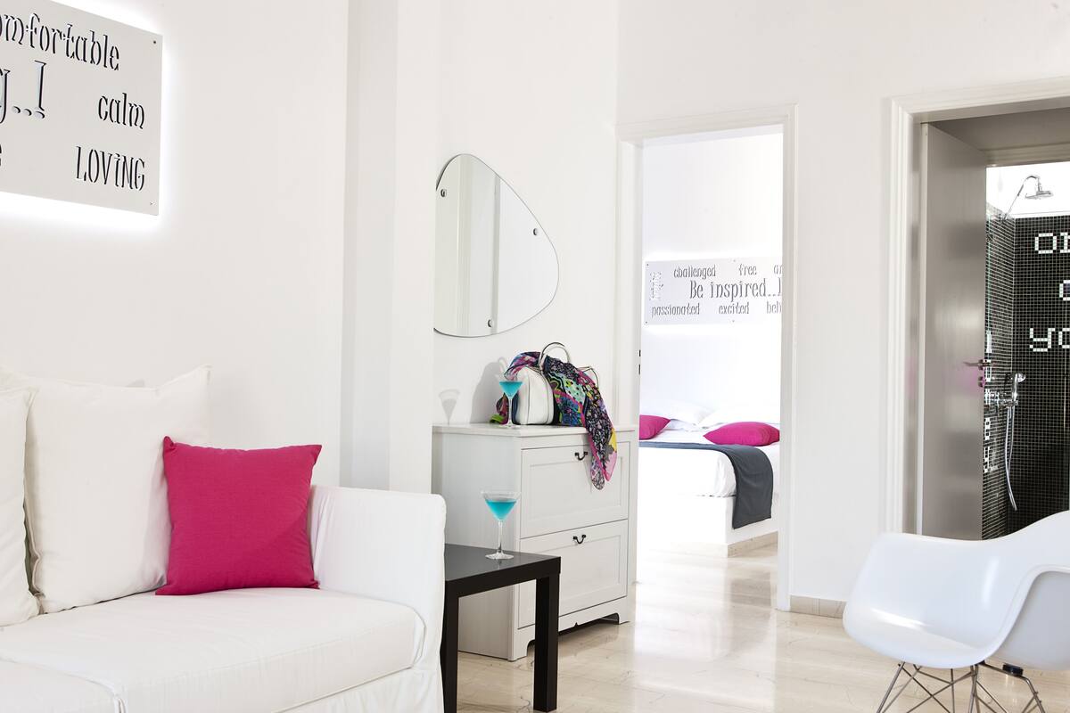 Une chambre d’hôtel moderne avec un canapé blanc, une petite table avec une boisson, un miroir et un lit avec des oreillers roses.