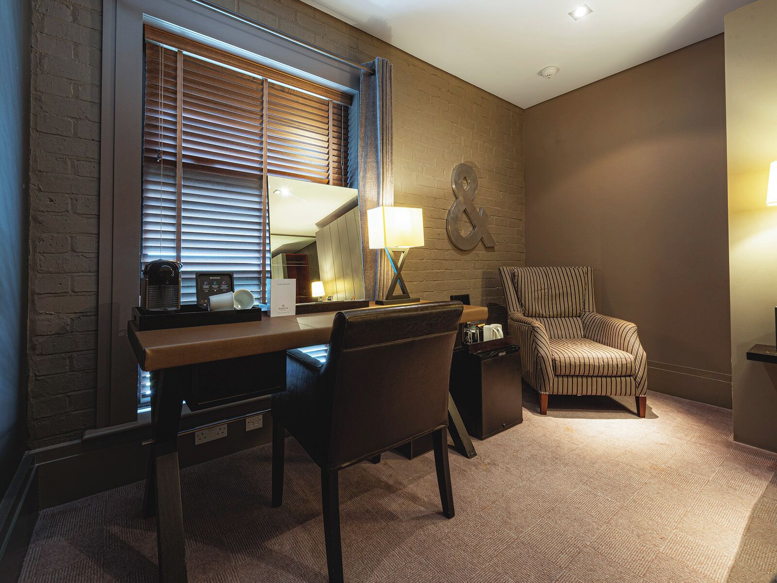 Ein Hotelzimmer mit Schreibtisch, Stuhl, Lampe, Spiegel und einer Wand mit dekorativem Metallschild.