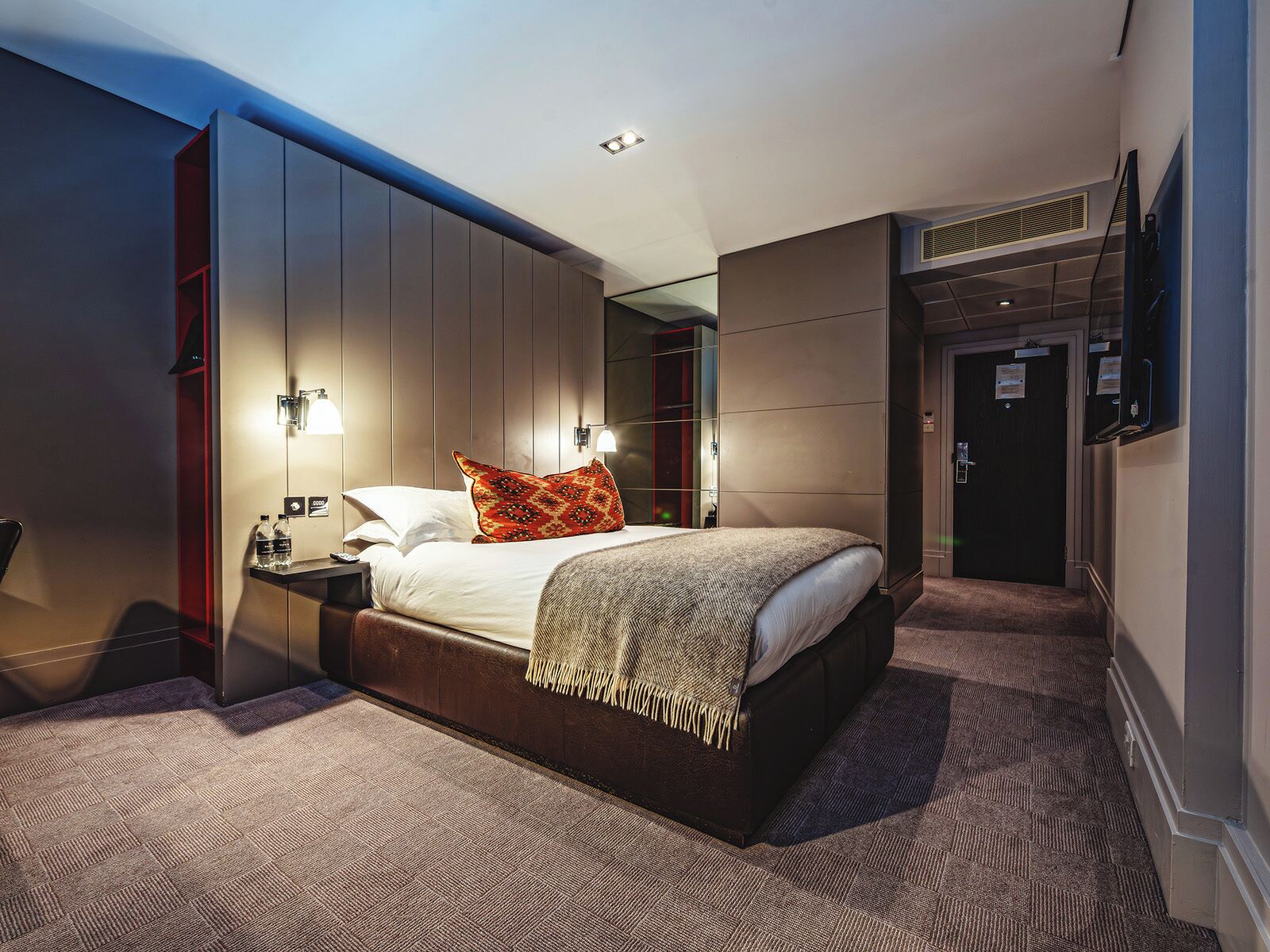 Quarto de hotel moderno com uma cama grande, armário embutido e uma pequena mesa de cabeceira.