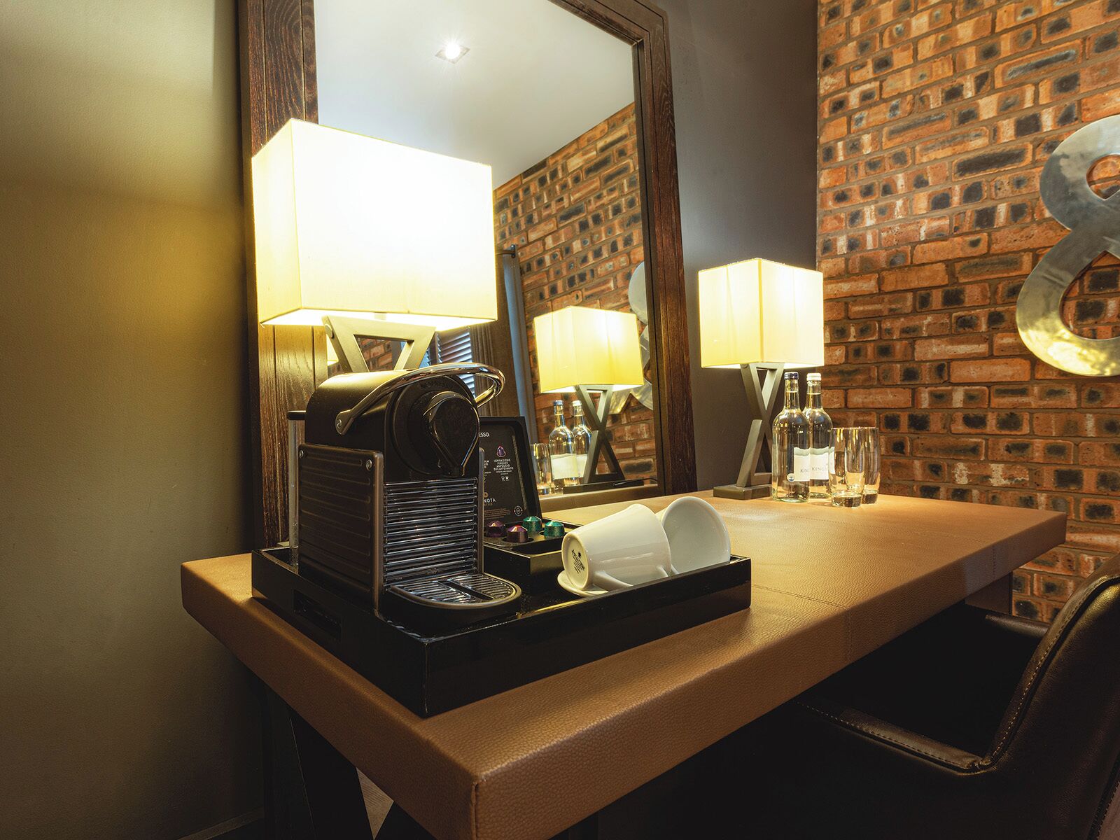 A hotel room with a coffee machine, a tray with cups, a mirror, and a brick wall.