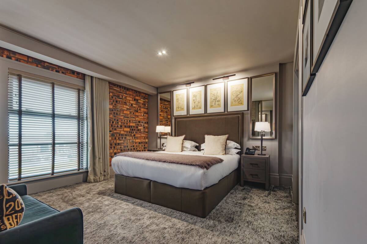 Une chambre d’hôtel avec un grand lit, un mur décoratif en briques, une fenêtre avec des stores, une table de chevet avec une lampe et des œuvres d’art encadrées accrochées au mur.