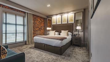 Habitación de hotel con una cama grande, pared decorada con ladrillos, ventana con persianas, mesita de noche con lámpara y obras de arte enmarcadas en la pared.