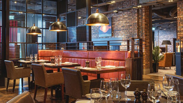A restaurant interior with brick walls, wooden slats, leather seating, and pendant lighting. - Dakota Edinburgh (South Queensferry)