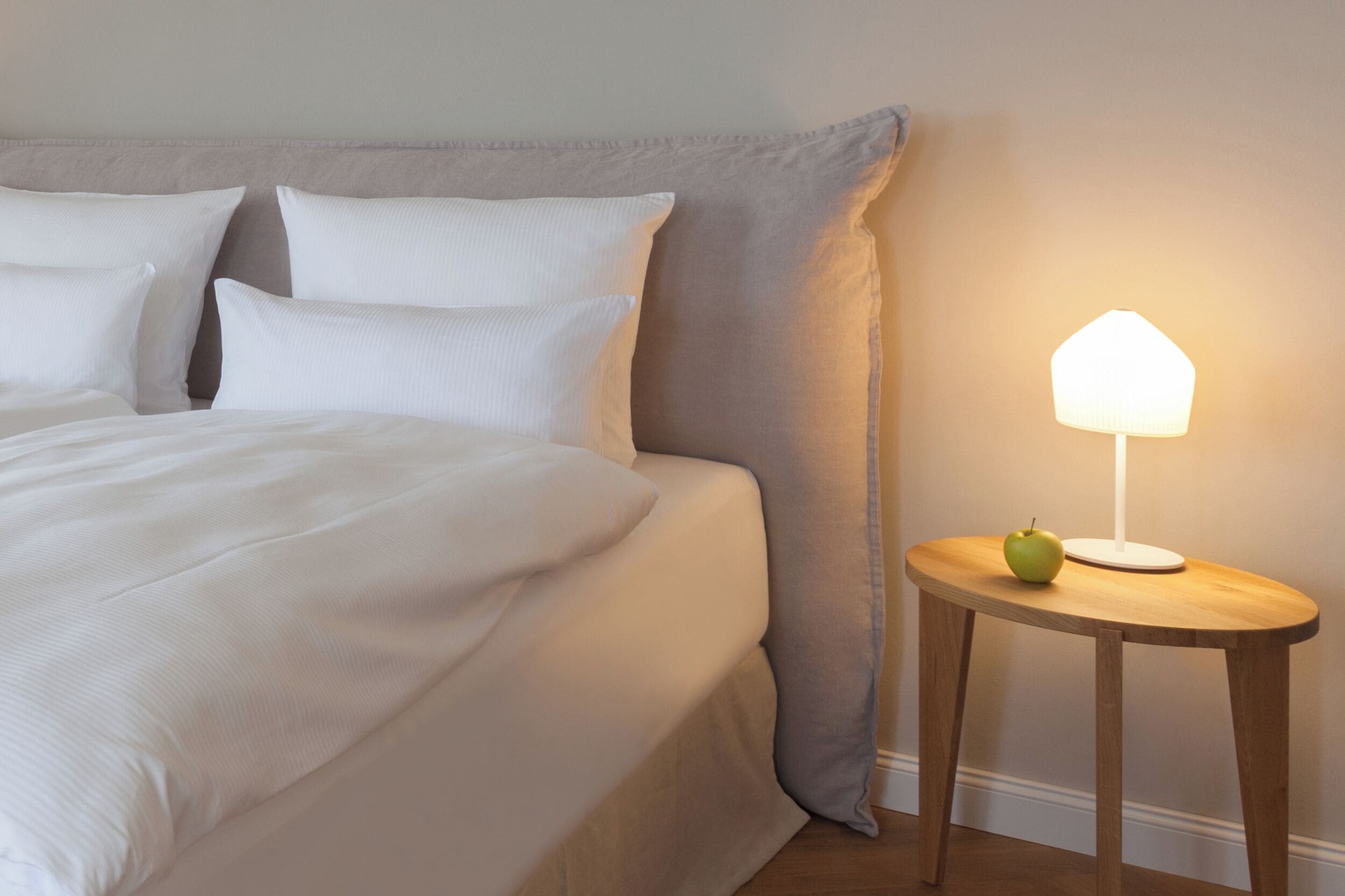 A neatly made bed with white linens and pillows, a round wooden nightstand with a lamp and an apple, and a large headboard against a plain wall.