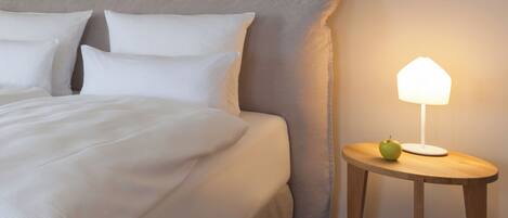 A neatly made bed with white linens and pillows, a round wooden nightstand with a lamp and an apple, and a large headboard against a plain wall.