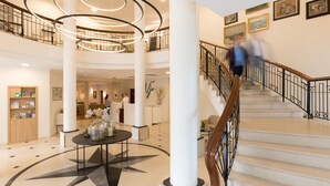 A spacious lobby with a central circular light fixture, a staircase with a wooden handrail, and a reception desk with framed artwork on the walls. - Vju Hotel Rügen (Göhren)