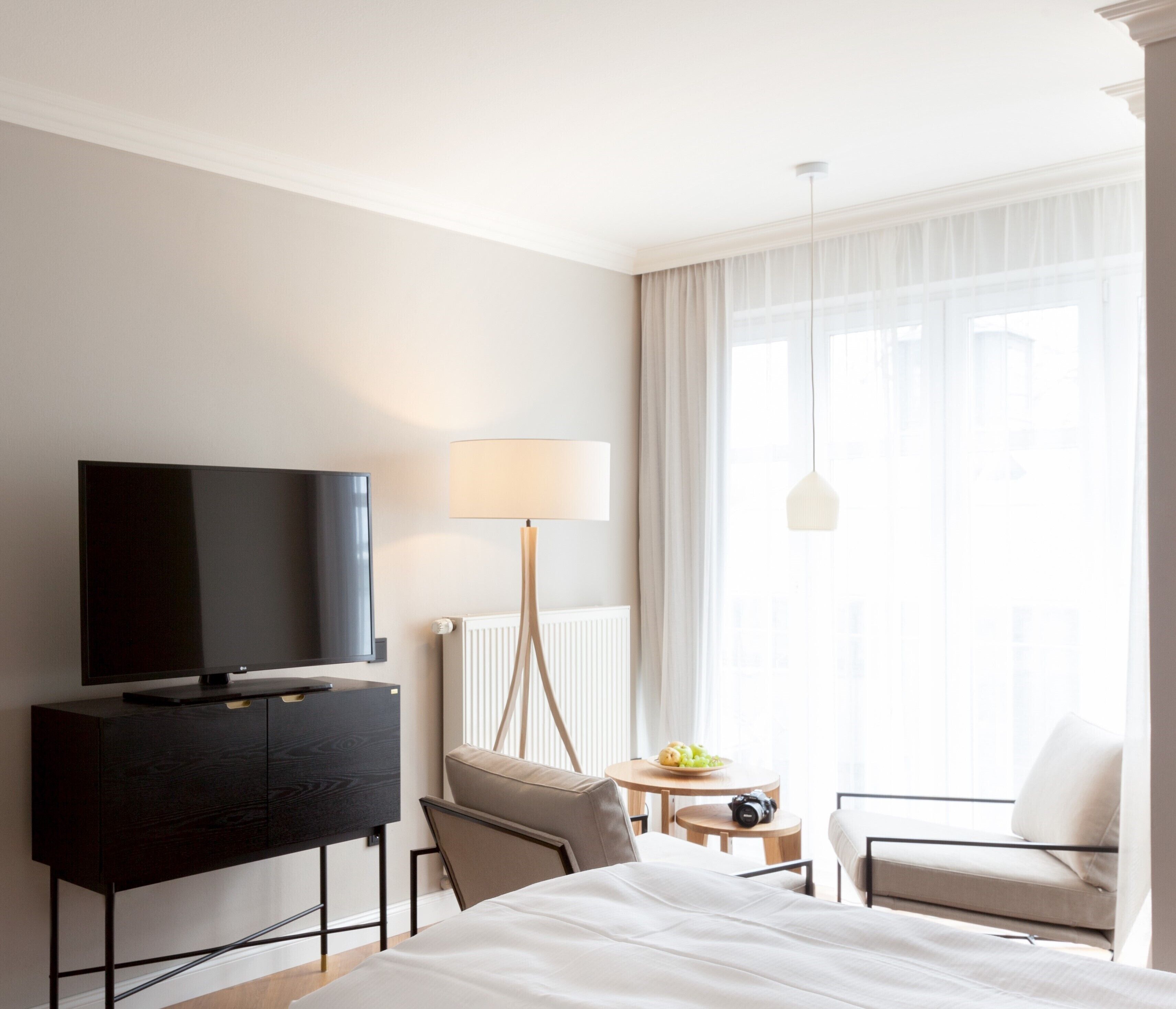 Habitación de hotel con cama, televisión sobre una cómoda de madera oscura, lámpara, silla, mesita con frutas y ventana con cortinas.