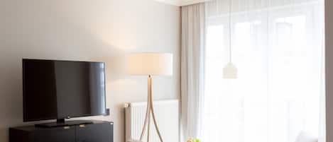 A hotel room with a bed, a TV on a dark wooden dresser, a lamp, a chair, a small table with fruits, and a window with curtains.
