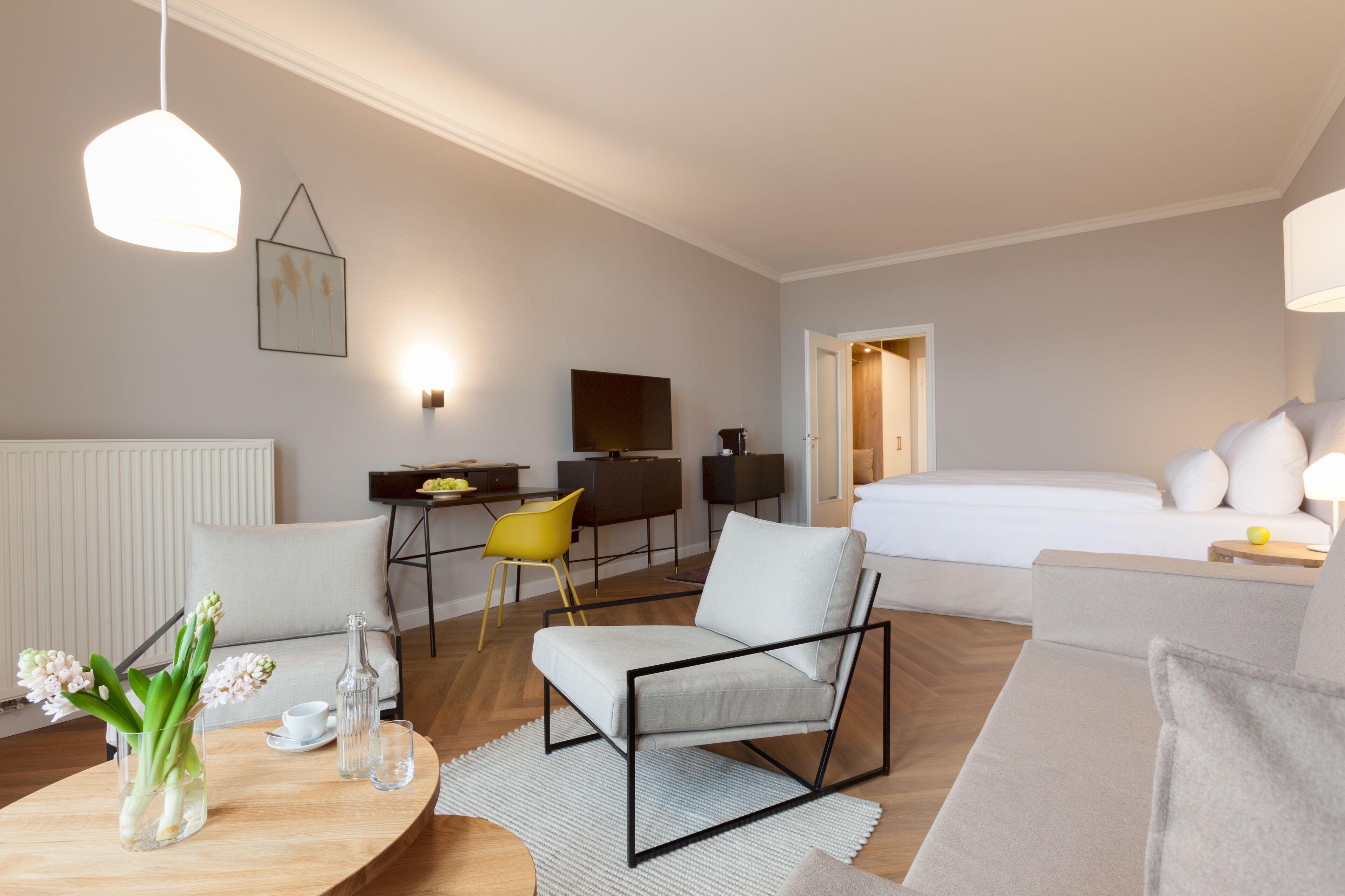 A modern hotel room with a bed, a sofa, a dining table with flowers, a TV, and a desk.