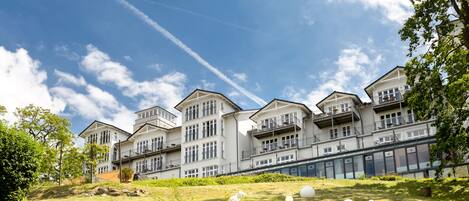 A row of white multi-story buildings with balconies, set against a clear blue sky with some clouds.