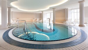 A large indoor swimming pool with a tiled floor and a central staircase. - Vju Hotel Rügen (Göhren)