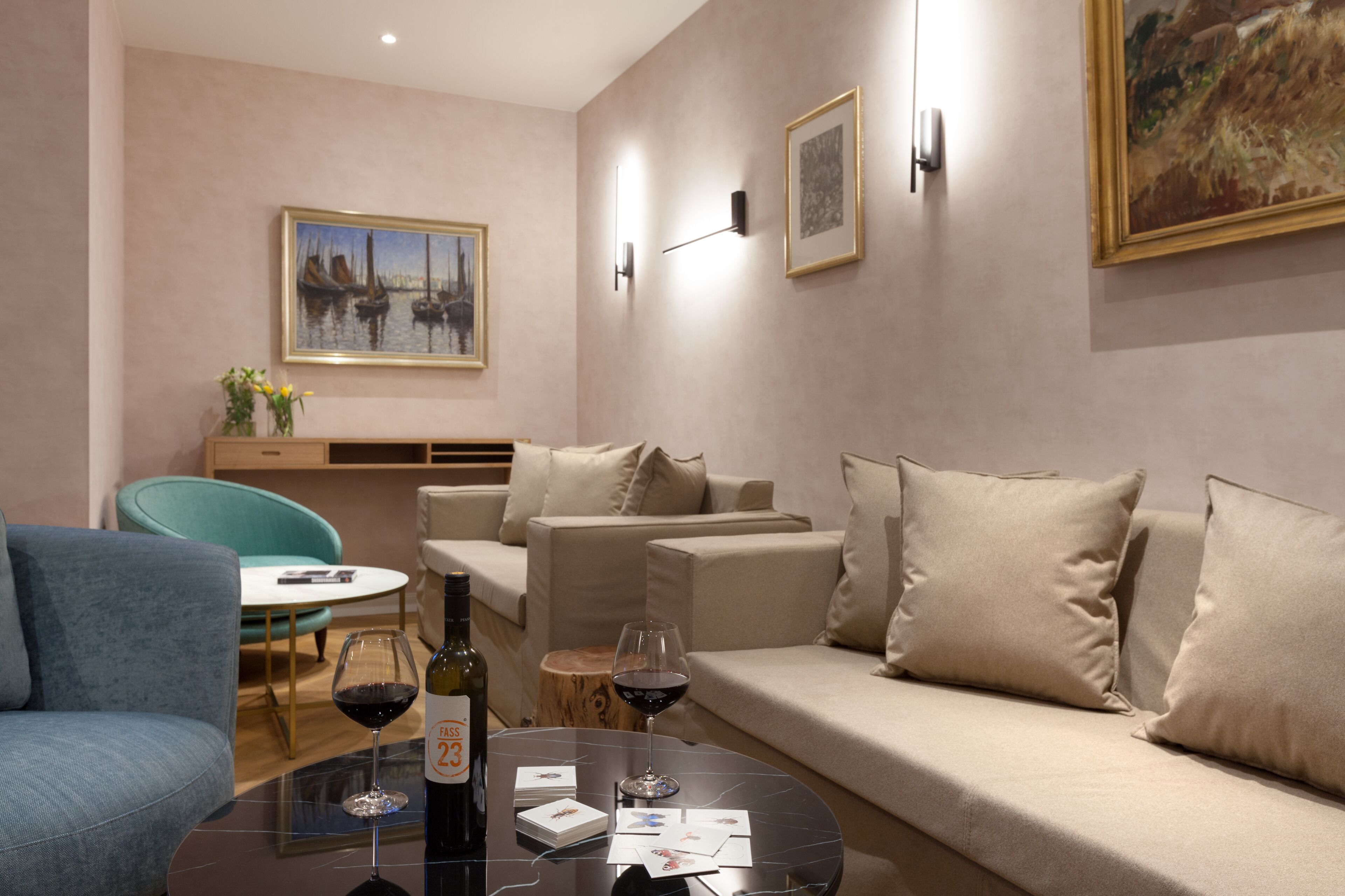 A modern living room with a sofa, a coffee table with wine and cards, a teal chair, and a painting on the wall.