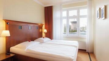 Ein Hotelzimmer mit Doppelbett, zwei Nachttischlampen, einem Holzkopfstück, einem Fenster mit Vorhängen und einer Heizung.