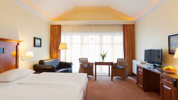 A hotel room with a large bed, a desk with a television, two chairs, a small table with a vase, and a window with curtains.