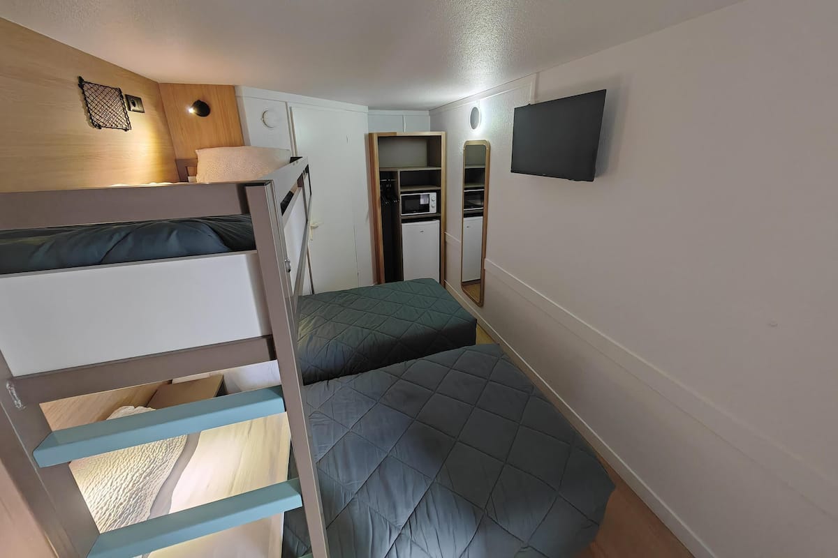 Een compacte kamer met stapelbedden, een klein keukentje, een spiegel en een aan de muur bevestigde televisie.
