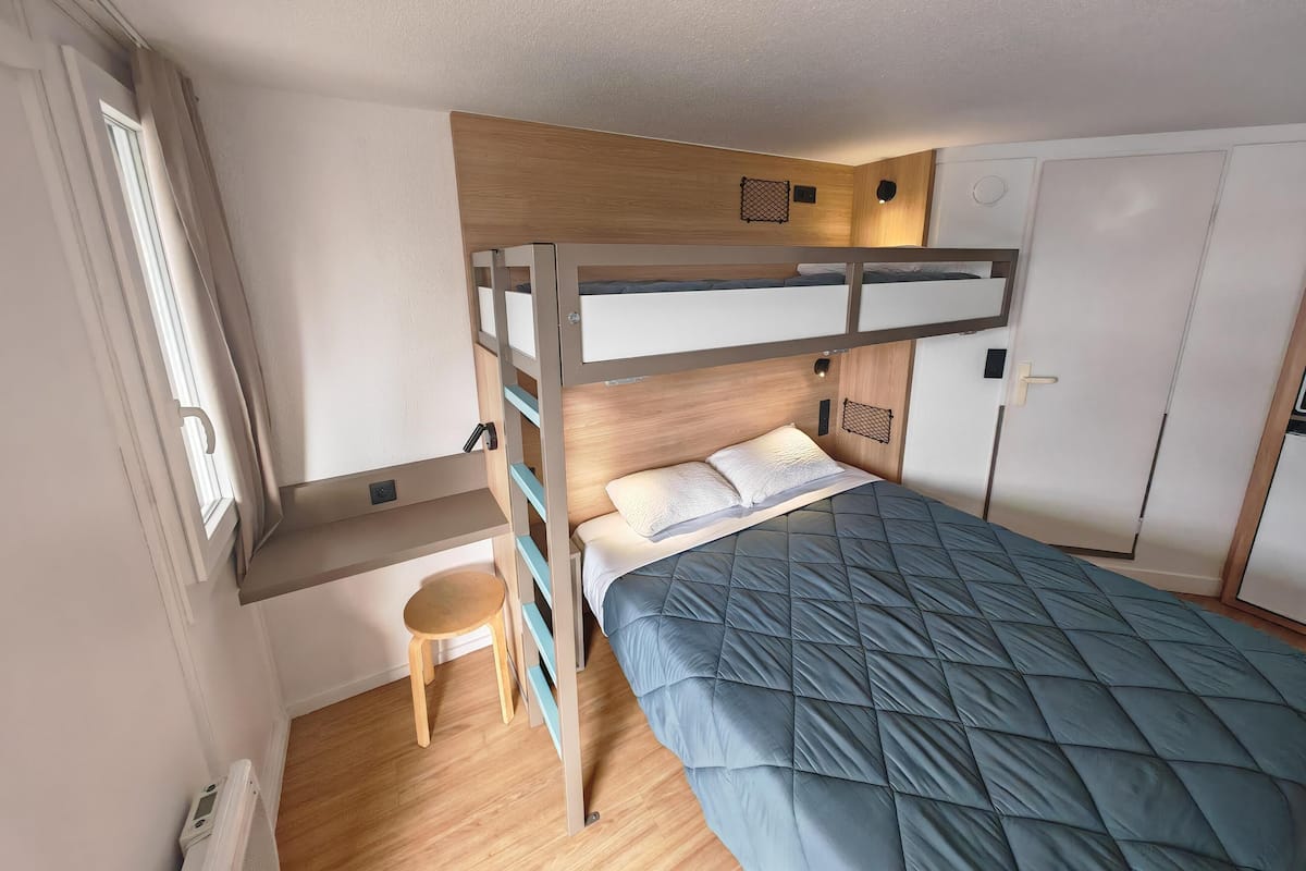 Een slaapkamer met een stapelbed, een bureau en stoel, een kledingkast en een klein krukje.