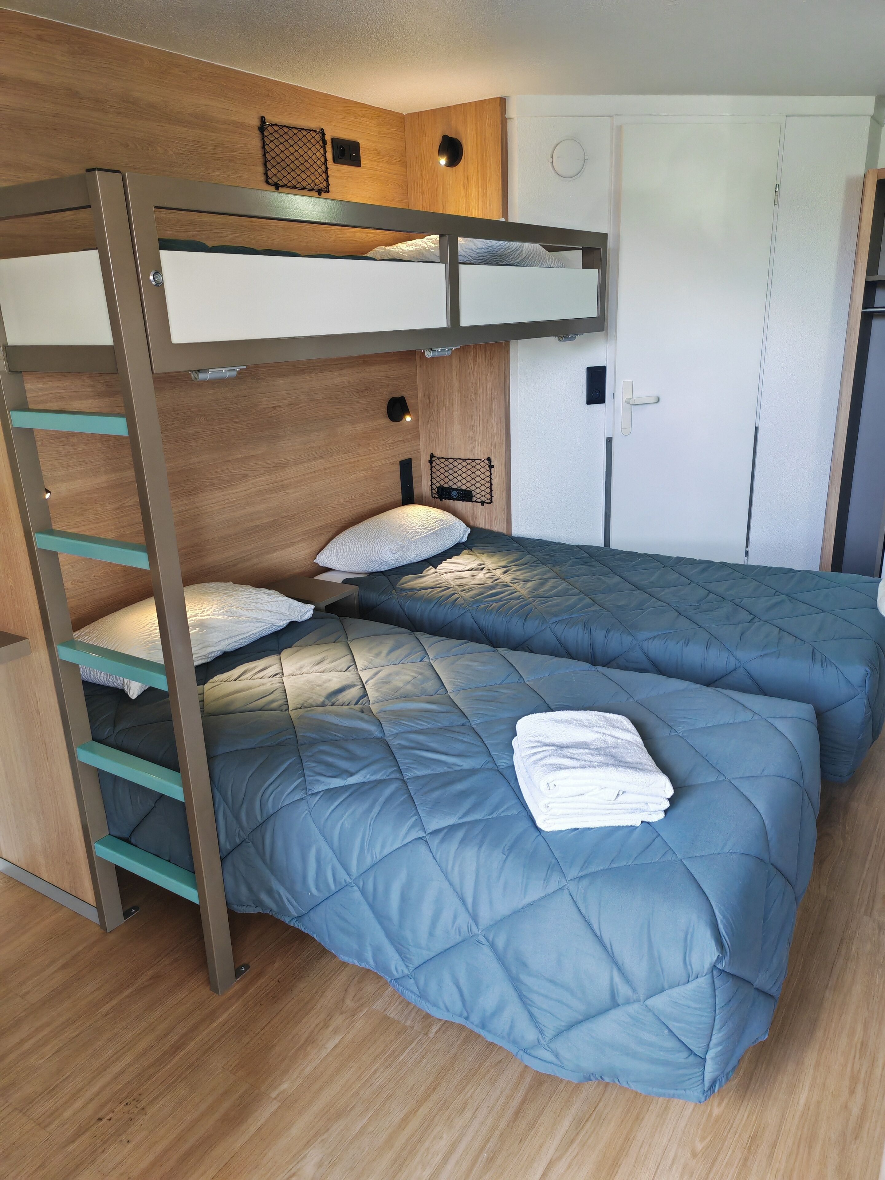 Habitación con literas, una escalera de madera, dos camas con ropa de cama azul, una puerta blanca y un estante pequeño.