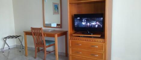 A room with a wooden TV stand, a flat-screen TV, a wooden desk, a chair, and a mirror.