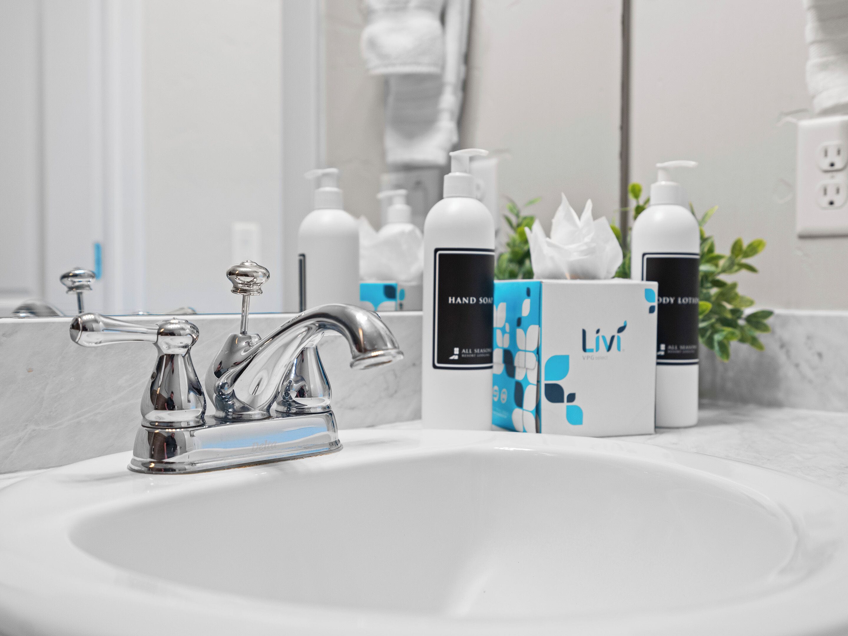 Ein Badezimmerwaschbecken mit einem modernen Wasserhahn, einem Spiegel und Pflegeprodukten wie Handseife und Körperlotion.