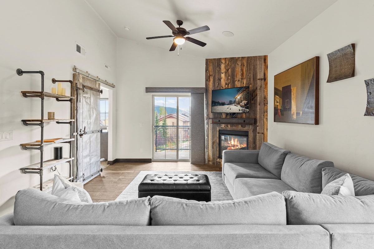 A spacious living room with a grey sofa, a fireplace, a mounted TV, and a ceiling fan.