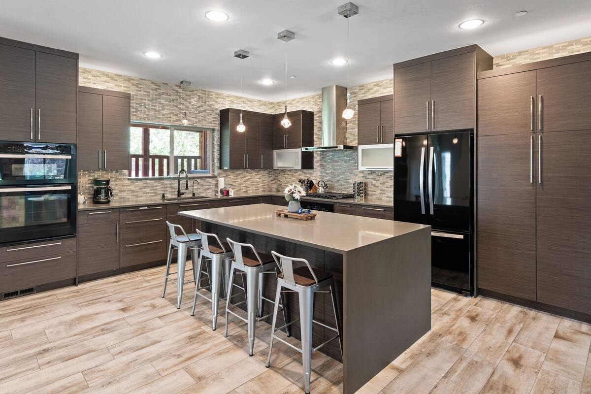 A modern kitchen with dark wood cabinets, a large island with bar stools, stainless steel appliances, and a brick backsplash.