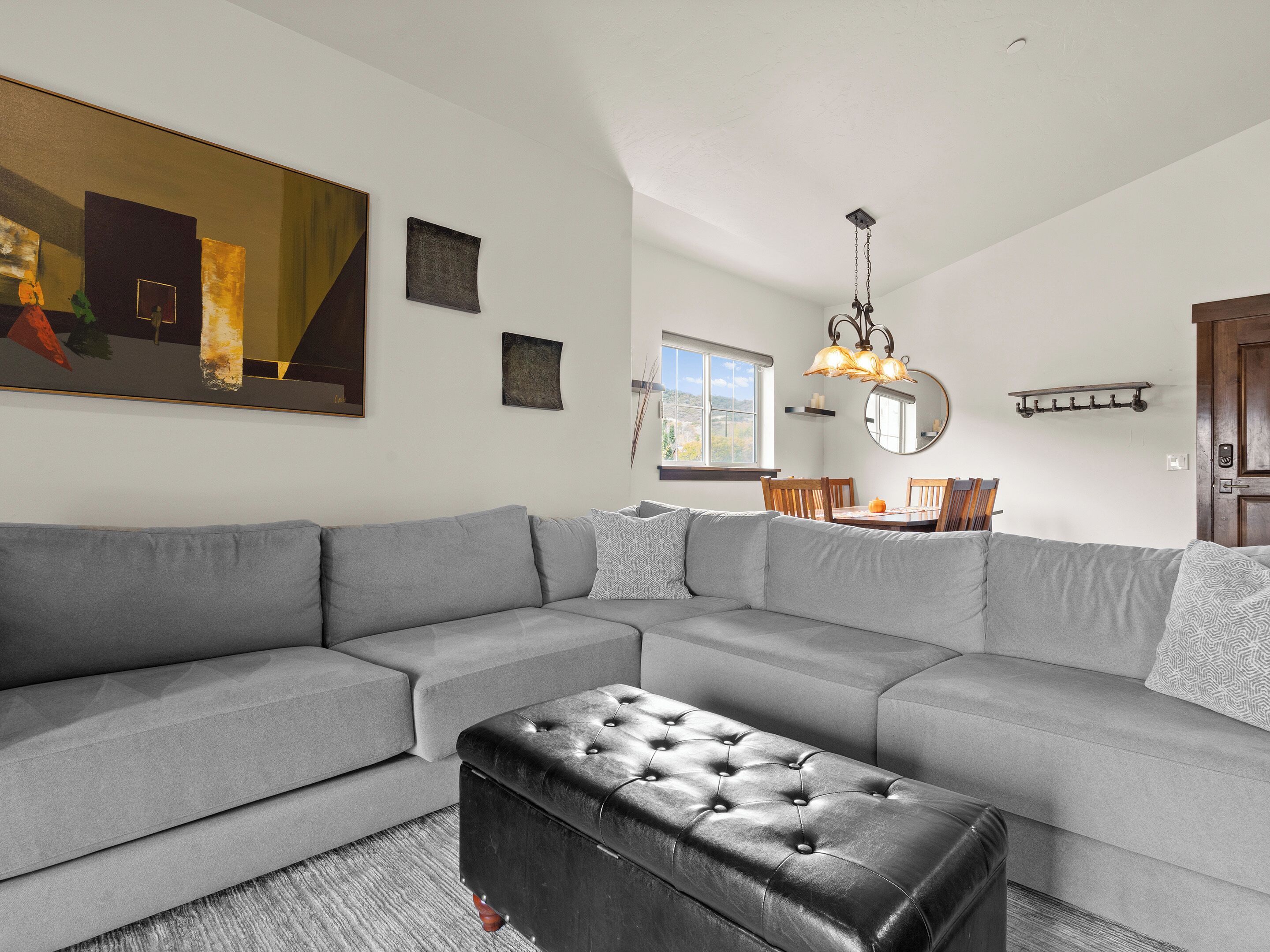 A spacious living room with a grey sectional sofa, a black leather ottoman, and a dining area with a wooden table and chairs.