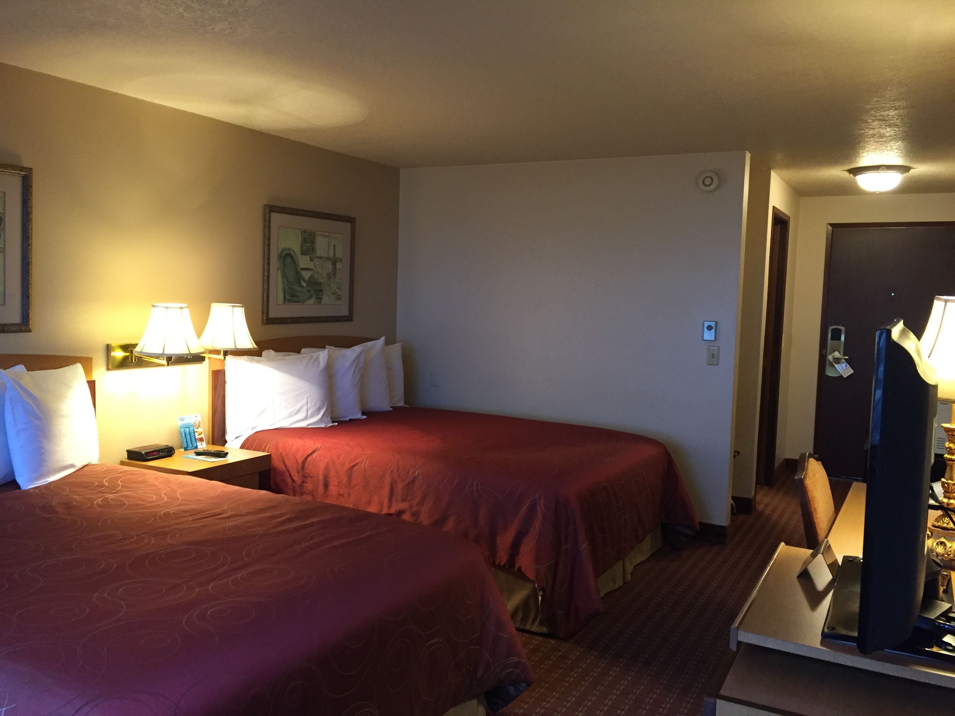 A hotel room with two beds, a nightstand, a desk with a lamp, and a door with a lock.