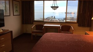 A hotel room with a bed, two chairs, a desk, a television, and a window with a view of a port and cranes. - Port Angeles Inn (Port Angeles)