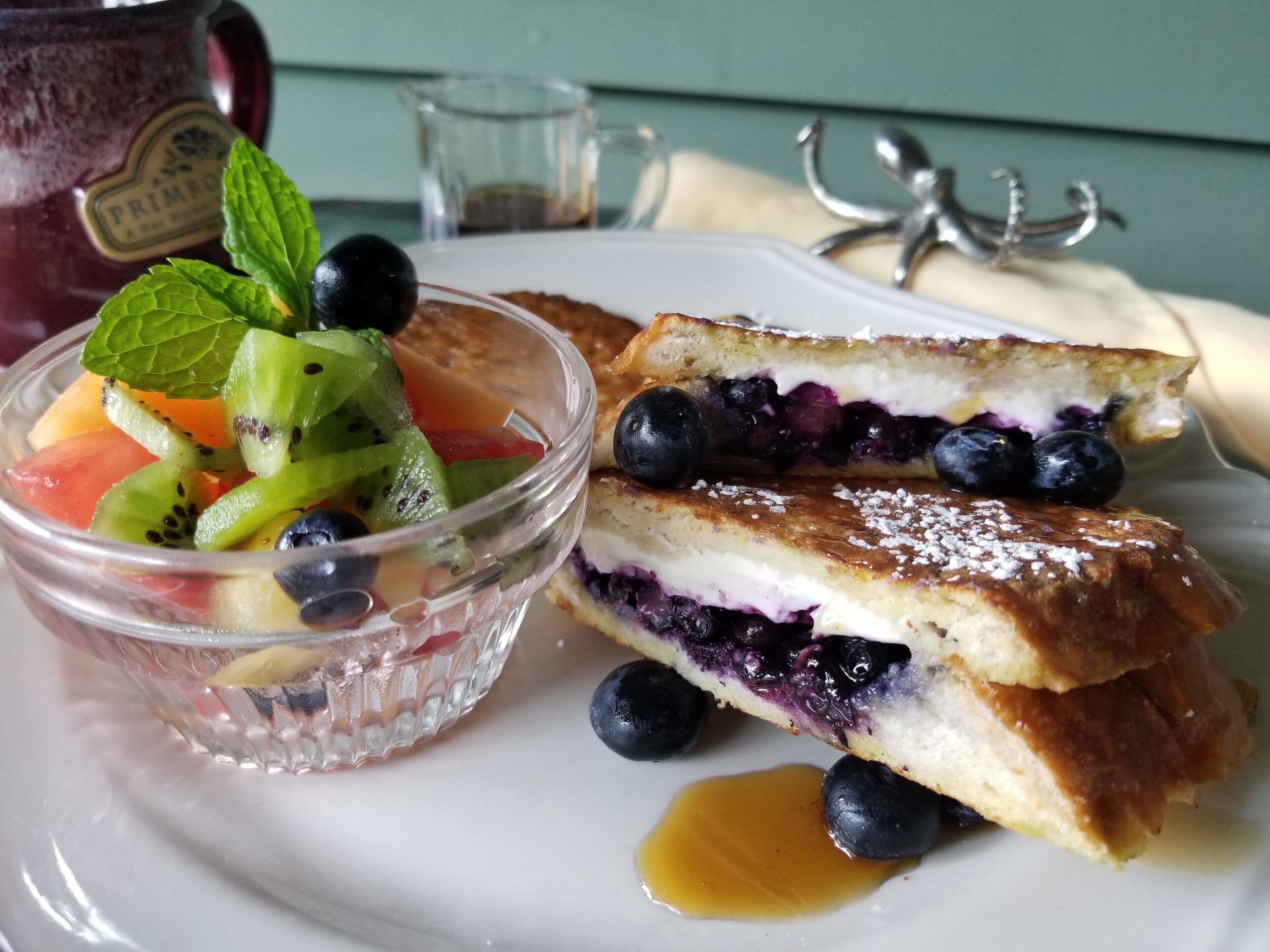 A dessert plate with a fruit salad, a slice of blueberry cake, and a small pitcher of syrup.