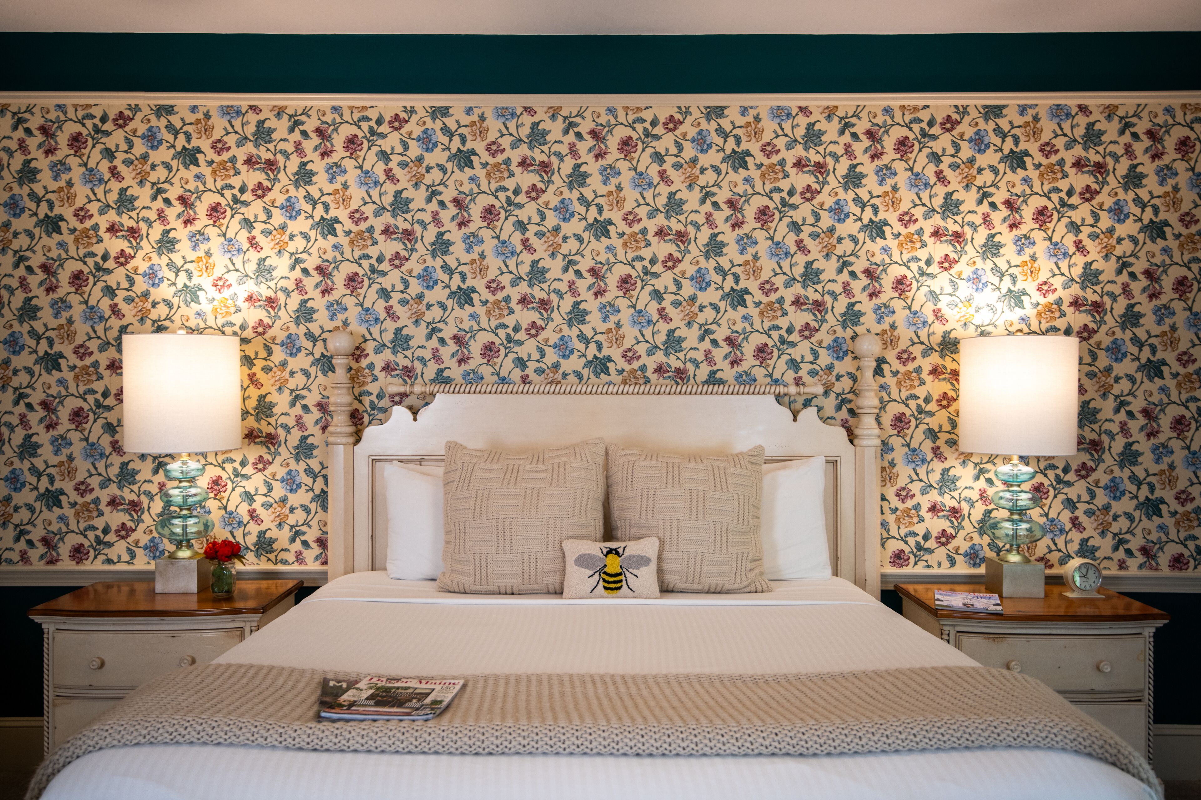 A neatly made bed with a white headboard, two bedside tables with lamps, and floral wallpaper.