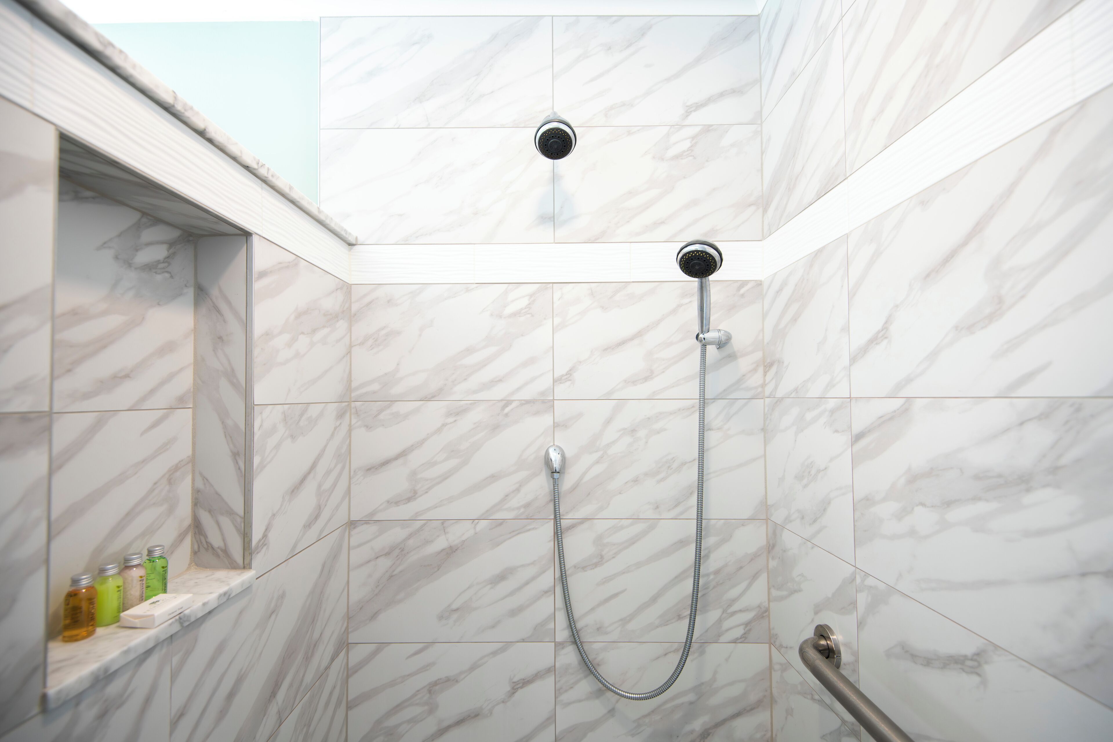 A shower area with a handheld showerhead and a fixed showerhead, marble tiles, and a shelf with toiletries.