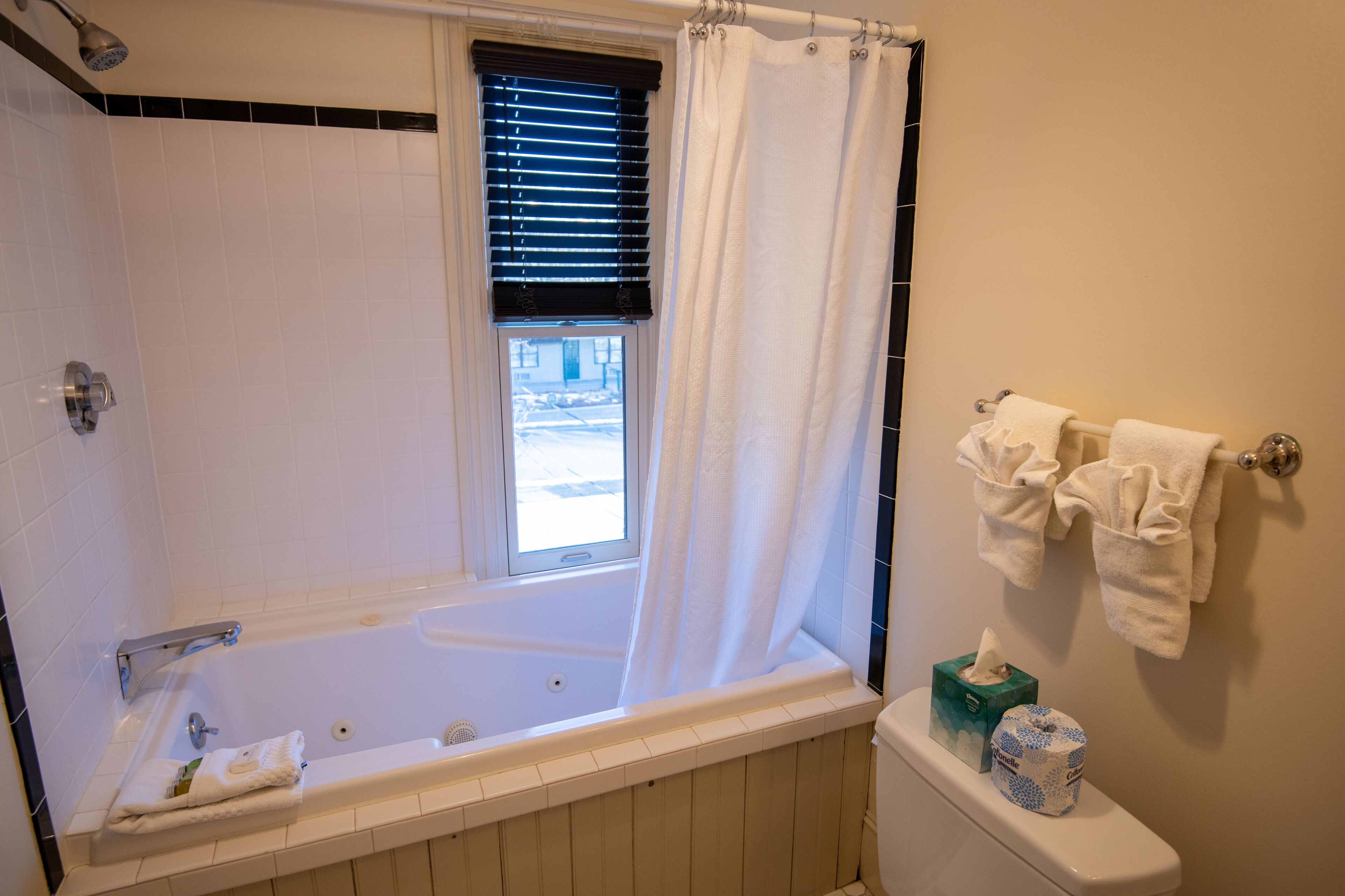 Een badkamer met een bad, een toilet, een raam met zonwering en een handdoekrek met handdoeken.